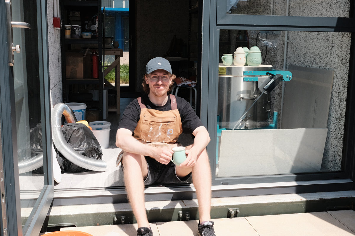 Person sitting on a step holding a mug, wearing an apron, with a workshop in the background.