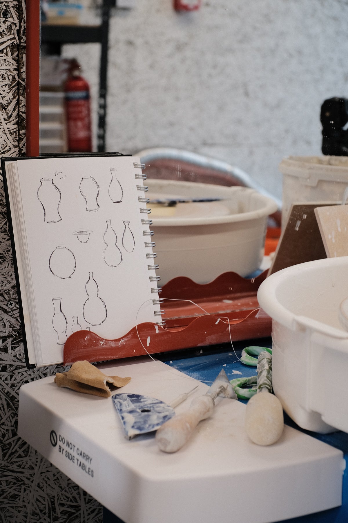 A white spiral-bound sketchbook with hand-drawn vase and vessel outlines sits on a workbench surrounded by pottery tools, a wooden paddle, a small tube of clay slip, and white ceramic bowls in a workshop setting.